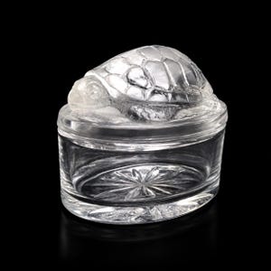 Può includere: Un portagioie in vetro trasparente con un coperchio caratterizzato da una statuetta di tartaruga scolpita sulla parte superiore. La base del contenitore è cilindrica con un motivo a stella inciso sul fondo, mentre il coperchio è liscio.