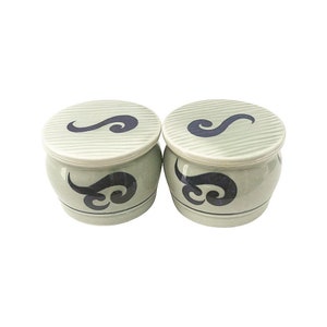Op de afbeelding: Twee keramische containers met deksels, elk met een gestileerd blauw swirl-ontwerp op een lichtgroene achtergrond. De containers hebben een afgeronde vorm, die doet denken aan kleine vaten.
