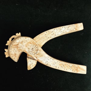 May include: A wooden nutcracker carved in the shape of a bird's head with intricate details. The nutcracker is made of light brown wood and has a geometric pattern on the handles.