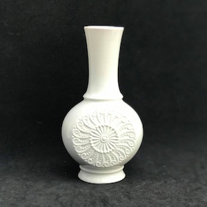 Może przedstawiać: Biały ceramiczny wazon z wytłoczonym wzorem kwiatowym. Wazon ma okrągłe dno i wysoką, smukłą szyjkę.