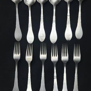 May include: A collection of vintage silverware, including six spoons and six forks, arranged on a black background. The spoons have oval bowls and decorative handles. The forks have four tines and matching handles. The silverware appears to be made of silver.