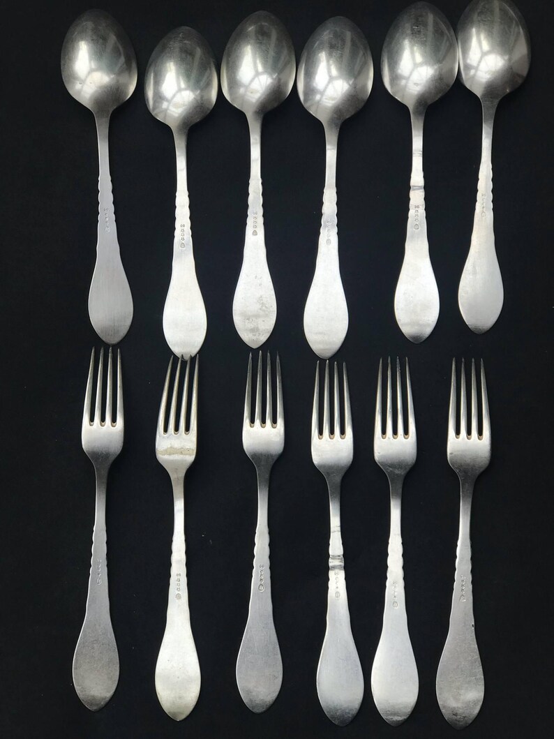 May include: A collection of vintage silverware, including six spoons and six forks, arranged on a black background. The spoons have oval bowls and decorative handles. The forks have four tines and matching handles. The silverware appears to be made of silver.