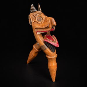 May include: A carved wooden nutcracker shaped like a stylized animal head with pointed ears and a snout. The wood features natural brown tones with painted red and black accents on the mouth and tongue area, held together by a metal hinge pin.