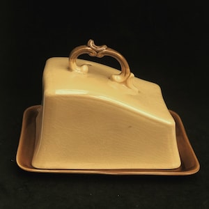 Puede incluir: Una cúpula de queso de cerámica con una base marrón y una tapa marrón claro. La tapa tiene un asa decorativa.