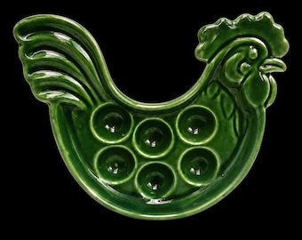 May include: A glossy, dark green ceramic egg tray shaped like a rooster. The tray features six circular indentations for holding eggs, with the rooster's head and tail forming the outer edges of the dish. The surface is smooth and reflective.