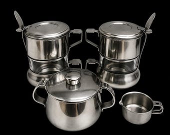 May include: A set of stainless steel kitchenware consisting of two stacked containers with lids and upright handles, a lidded sugar bowl with two side handles, and a small creamer pitcher with a single handle, all featuring a polished metallic finish.
