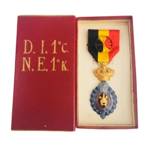Può includere: Una medaglia d'oro e argento con un designo in smalto blu e rosso. La medaglia è in una scatola rossa con lettere dorate che recitano "D. I. 1°c. N. E. 1°k."