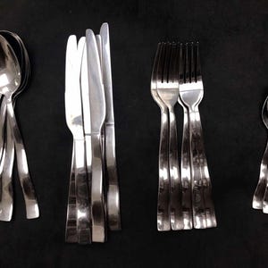 May include: A collection of stainless steel silverware, including spoons, knives, and forks, arranged on a black surface. The spoons have rounded bowls, the knives have serrated edges, and the forks have four tines. The silverware is clean and reflective.