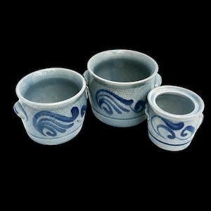 May include: Three light blue ceramic crocks of varying sizes, each featuring a dark blue hand-painted swirl pattern and two small side handles. The cylindrical vessels have a smooth, glazed finish and open tops, arranged in a cluster against a black background.