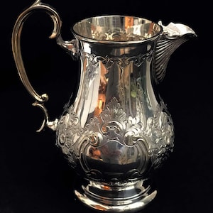 Puede incluir: Una jarra de plata con un diseño ornamentado y un asa curva. La jarra tiene una base ancha y un pico con un labio decorativo.