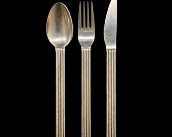 May include: A silver-toned metal flatware set consisting of a spoon, a four-tined fork, and a knife. Each piece features a long, straight handle with a distinct vertical ribbed or fluted texture running the entire length of the handle.