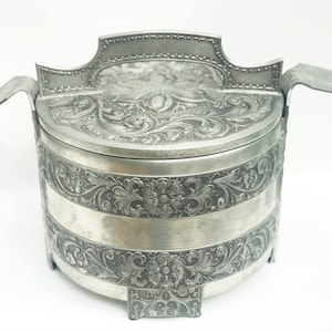Puede incluir: Un recipiente plateado antiguo con intrincados grabados florales y de volutas. El recipiente tiene un cuerpo redondeado, una tapa y dos asas. El diseño incluye una banda decorativa alrededor del centro y una parte superior detallada.
