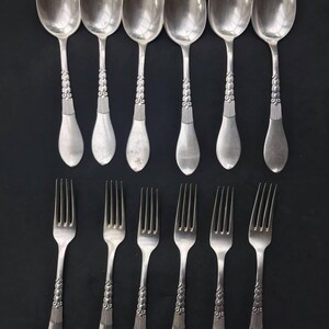 May include: A set of vintage silverware, including six spoons and six forks, arranged on a black surface. The silverware is silver-colored with ornate detailing on the handles. The spoons have oval bowls, and the forks have four tines.