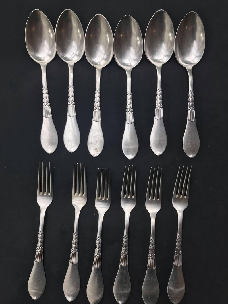 May include: A set of vintage silverware, including six spoons and six forks, arranged on a black surface. The silverware is silver-colored with ornate detailing on the handles. The spoons have oval bowls, and the forks have four tines.