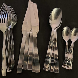 May include: A collection of stainless steel silverware, including forks, knives, and spoons, arranged on a dark surface. The forks have a wavy design, while the knives have serrated edges. The spoons are of two sizes.