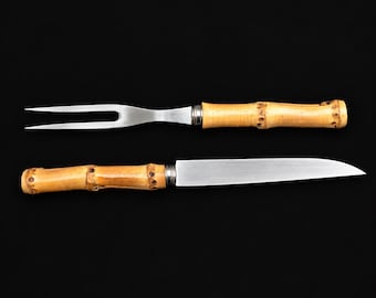 May include: A two-pronged carving fork and a straight-edged carving knife, both featuring natural light brown bamboo handles with visible nodes and silver-toned metal blades, positioned horizontally against a solid black background.