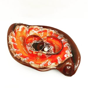 Peut inclure: Un plat en céramique de forme organique, avec un extérieur marron brillant et un intérieur orange et blanc vif. Deux boucles d'oreilles créoles argentées et un bracelet avec une perle reposent à l'intérieur du plat, créant une présentation élégante.