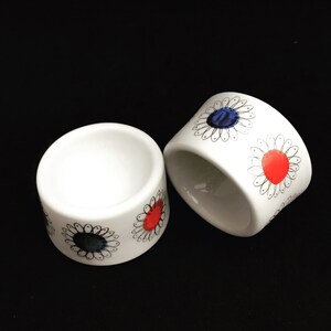 Puede incluir: Dos soportes para huevos de cerámica blanca con diseños florales en blanco y negro. Un soporte tiene una flor roja y azul, el otro tiene una flor azul.