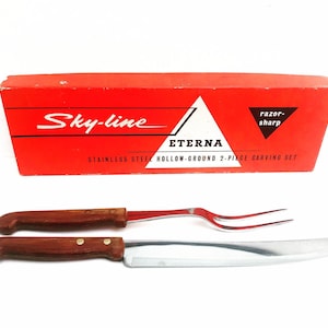 Peut inclure: Une boîte rouge avec le texte "Sky-line ETERNA STAINLESS STEEL HOLLOW-GROUND 2-PIECE CARVING SET" et un couteau et une fourchette à découper avec des manches en bois.