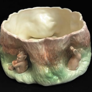 Op de afbeelding: Een keramische kom in de vorm van een boomstronk met twee konijnen aan de zijkanten. De kom is versierd met bruine, groene en witte glazuur.