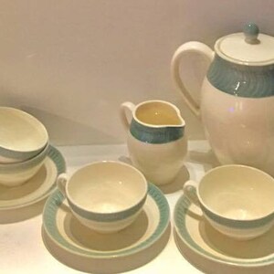 May include: A ceramic tea set featuring a cream-colored base with teal-colored bands and ribbed detailing. The set includes a teapot with a lid, a creamer, a lidded sugar bowl, and several teacups with matching saucers arranged on a flat surface.