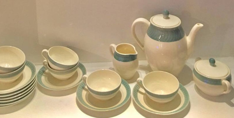 May include: A ceramic tea set featuring a cream-colored base with teal-colored bands and ribbed detailing. The set includes a teapot with a lid, a creamer, a lidded sugar bowl, and several teacups with matching saucers arranged on a flat surface.