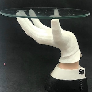 Puede incluir: Una mano de cerámica blanca con un guante blanco sostiene una bandeja de servir de vidrio transparente. La mano está unida a una base blanca y negra.