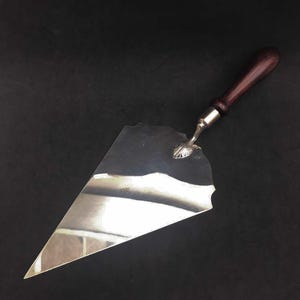 May include: A silver cake server with a wooden handle. The server has a scalloped edge and a shell-shaped design at the top of the handle.