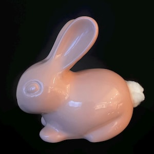 Może przedstawiać: Ceramiczna figurka królika w miękkim różowym odcieniu, z błyszczącym wykończeniem. Królik ma długie uszy, okrągły tułów i puszysty biały ogon. Figurka jest umieszczona na czarnym tle.