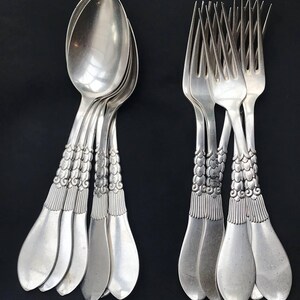 May include: A collection of silver spoons and forks arranged against a black background. The silverware features ornate, detailed handles with a repeating pattern. The spoons have a rounded bowl shape, while the forks have four tines.