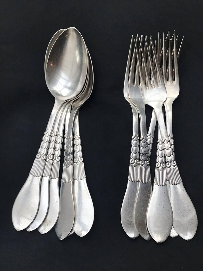 May include: A collection of silver spoons and forks arranged against a black background. The silverware features ornate, detailed handles with a repeating pattern. The spoons have a rounded bowl shape, while the forks have four tines.