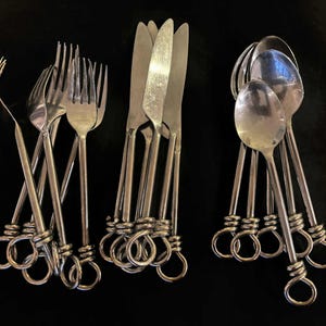 Może przedstawiać: Zestaw sztućców ze stali nierdzewnej, w tym widelce, noże i łyżki. Uchwyty mają unikalny pętelkowy design. Sztućce są ułożone na czarnej powierzchni, tworząc uderzający kontrast z błyszczącym metalem.