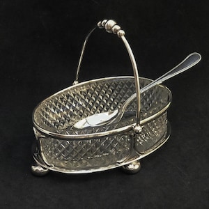 Op de afbeelding: Een zilveren suikerpot met een glazen inzet en een bijpassende lepel. De kom heeft een ruitvormig patroon en een handvat.