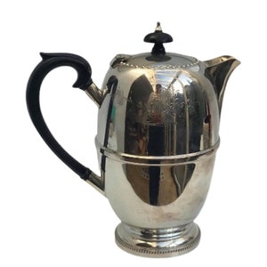 Op de afbeelding: Een zilveren theepot met een zwarte handgreep en een decoratief ontwerp. De theepot heeft een rond lichaam en een klein deksel met een knop bovenop.