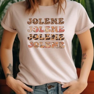 May include: A light beige short-sleeved t-shirt featuring the name JOLENE printed four times in a stacked design. The text alternates between a brown and black leopard print pattern and a multi-colored floral pattern in shades of orange and pink.