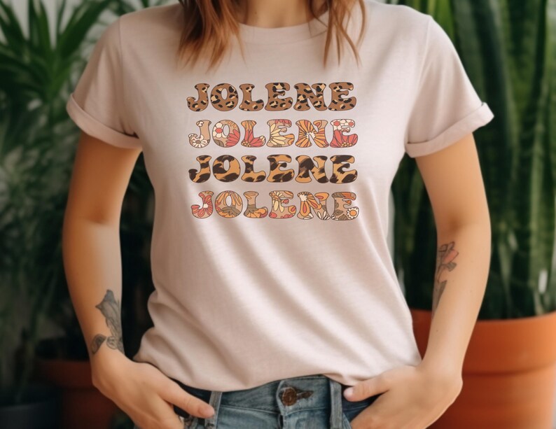 May include: A light beige short-sleeved t-shirt featuring the name JOLENE printed four times in a stacked design. The text alternates between a brown and black leopard print pattern and a multi-colored floral pattern in shades of orange and pink.