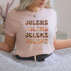 May include: A light pink short-sleeve t-shirt featuring the name JOLENE printed four times in a stacked design. Each line of text displays a different pattern, including leopard print, orange and white floral motifs, and brown animal spots.