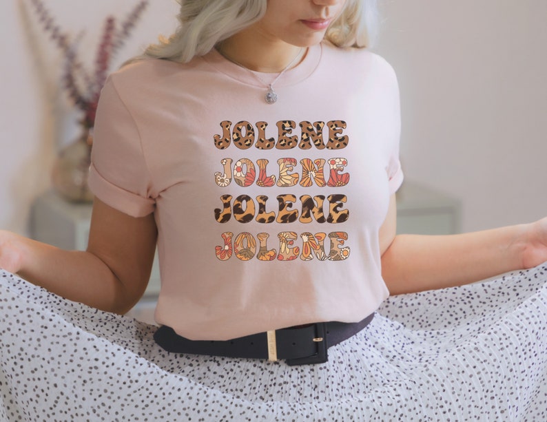 May include: A light pink short-sleeve t-shirt featuring the name JOLENE printed four times in a stacked design. Each line of text displays a different pattern, including leopard print, orange and white floral motifs, and brown animal spots.