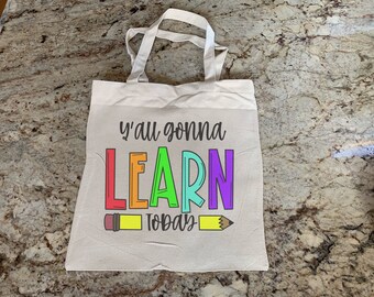 Può includere: Una borsa di tela bianca con il testo "Y'all gonna LEARN Today" in colori arcobaleno. Il testo è circondato da due matite gialle.