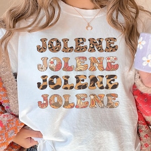 May include: A white t-shirt featuring the name JOLENE printed four times in a stacked design. The first and third lines have a brown leopard print pattern, while the second and fourth lines feature a colorful floral pattern in orange, red, and white.
