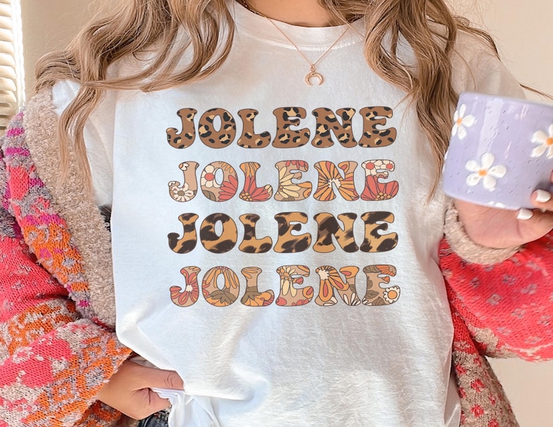 May include: A white t-shirt featuring the name JOLENE printed four times in a stacked design. The first and third lines have a brown leopard print pattern, while the second and fourth lines feature a colorful floral pattern in orange, red, and white.
