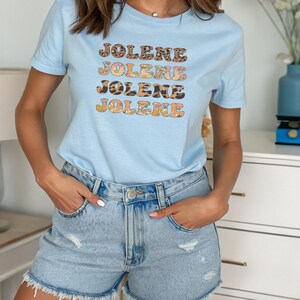 May include: A light blue short-sleeve t-shirt featuring the name JOLENE printed four times in a stacked design. Each line of text displays a different pattern, including leopard print, floral motifs, and solid colors, with a crew neckline.