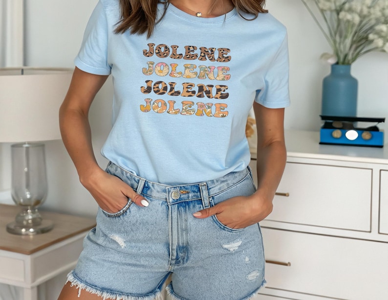 May include: A light blue short-sleeve t-shirt featuring the name JOLENE printed four times in a stacked design. Each line of text displays a different pattern, including leopard print, floral motifs, and solid colors, with a crew neckline.