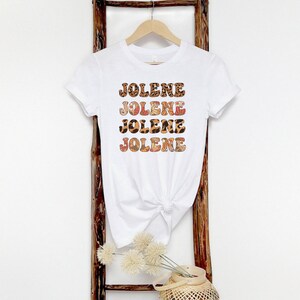 May include: A white short-sleeved t-shirt featuring the name JOLENE printed four times in a stacked design. The letters have a patterned fill with alternating leopard print and floral motifs in shades of brown, orange, and cream.