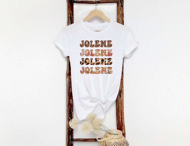 May include: A white short-sleeved t-shirt featuring the name JOLENE printed four times in a stacked design. The letters have a patterned fill with alternating leopard print and floral motifs in shades of brown, orange, and cream.