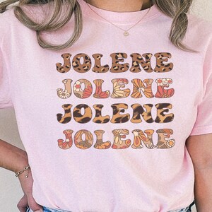 May include: A light pink short-sleeved t-shirt featuring the name JOLENE printed four times in a stacked design. The text alternates between a brown and black leopard print pattern and a multi-colored floral pattern in shades of orange, red, and cream.
