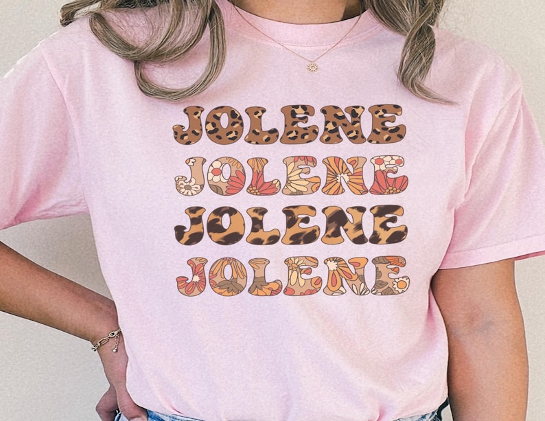 May include: A light pink short-sleeved t-shirt featuring the name JOLENE printed four times in a stacked design. The text alternates between a brown and black leopard print pattern and a multi-colored floral pattern in shades of orange, red, and cream.