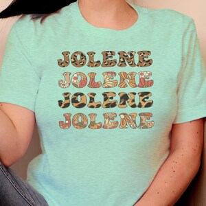 May include: A light mint green short-sleeve t-shirt featuring the name JOLENE printed four times in a stacked design. The first and third lines have a brown leopard print pattern, while the second and fourth lines feature a floral pattern.