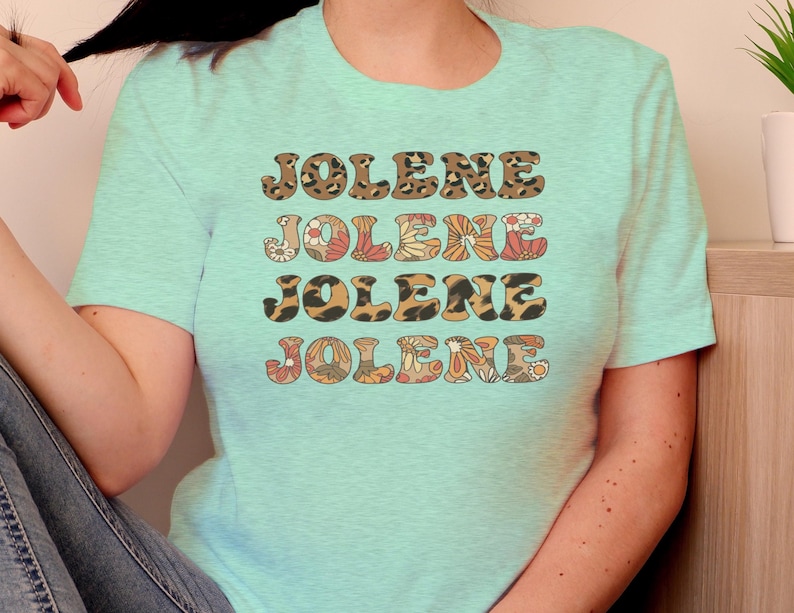 May include: A light mint green short-sleeve t-shirt featuring the name JOLENE printed four times in a stacked design. The first and third lines have a brown leopard print pattern, while the second and fourth lines feature a floral pattern.