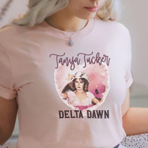 May include: Light pink t-shirt featuring a portrait of Tanya Tucker inside a heart-shaped design. The text "Tanya Tucker" is above the portrait, and "DELTA DAWN" is below. The shirt has short sleeves and a crew neck.